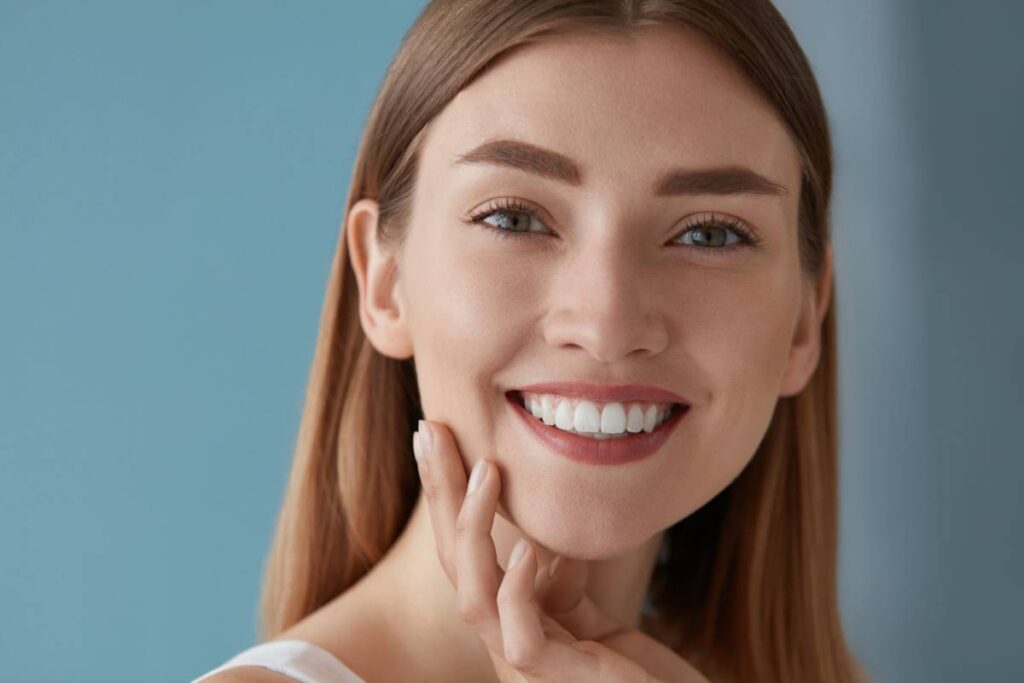 Smiling woman with long brown hair and flawless skin touches her face. She has bright eyes and white teeth, conveying happiness, set against a soft blue background.
