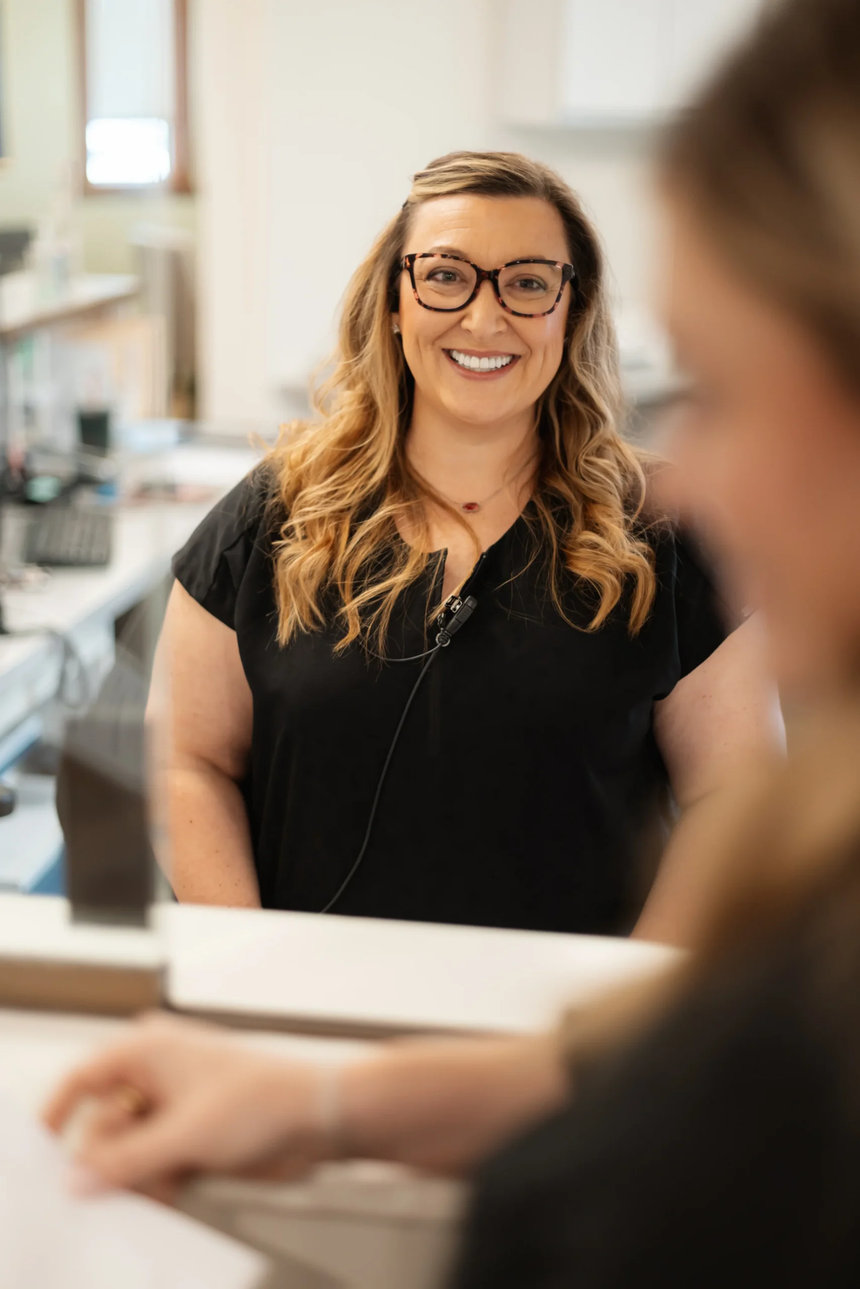 A woman with long, wavy hair and glasses smiles warmly at the camera in a softly lit office setting, conveying a friendly and inviting atmosphere.