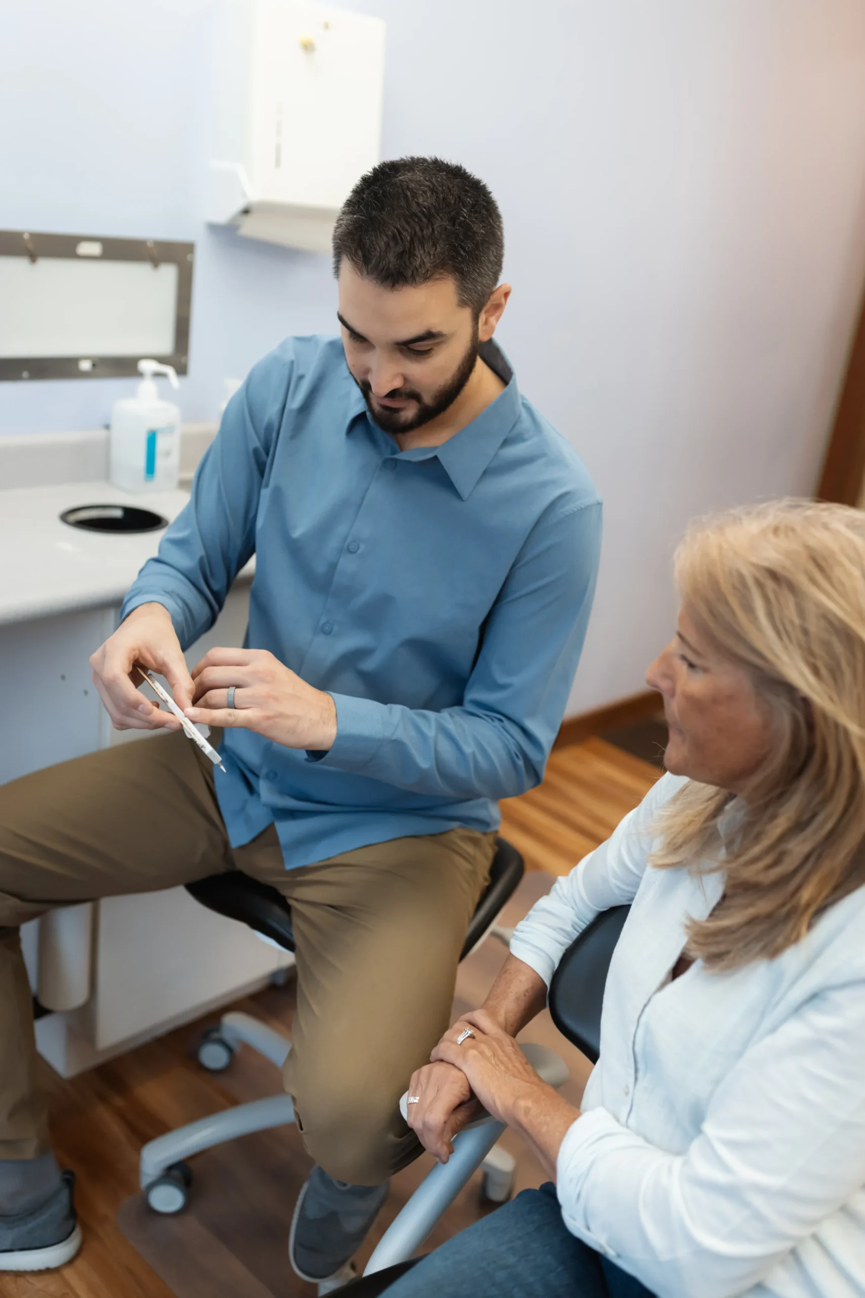 A man in a blue shirt explains a document to a woman in a white top, both seated in a clinical setting, conveying a professional and informative tone.