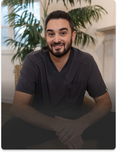A man with a beard and short hair, wearing a dark scrub top, sits smiling in a chair. He is in a room with large windows and lush green plants.
