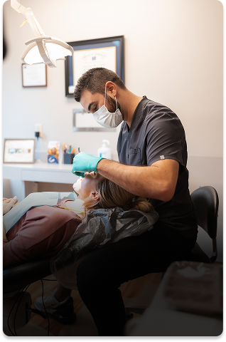 A dentist wearing a mask and gloves examines a patient lying in a dental chair. The setting is clinical and focused, conveying a sense of professionalism.