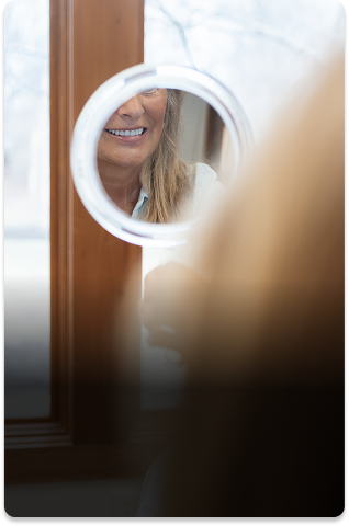 A woman’s face is partially reflected in a small round mirror, showing her smile. The background has a wooden window frame and a softly lit interior.