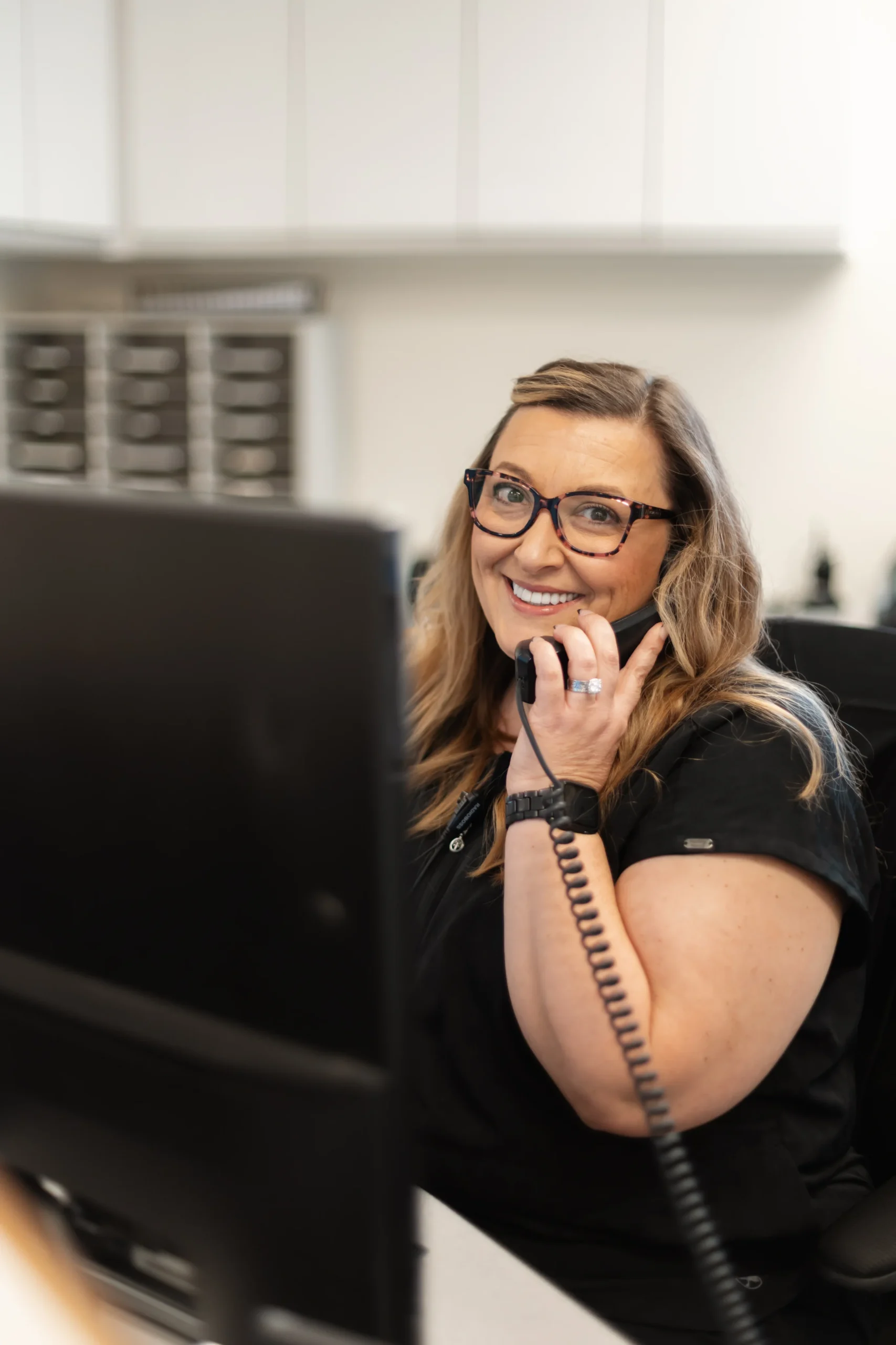 A woman with long hair and glasses smiling while on the phone at an office desk. She appears approachable and professional, conveying friendliness.