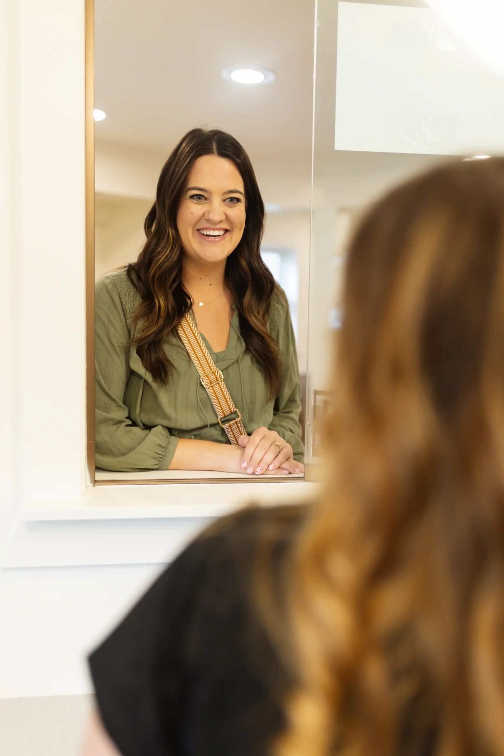 A woman with long brown hair and a green shirt smiles warmly while standing at a service counter. Another person is partially visible in the foreground.