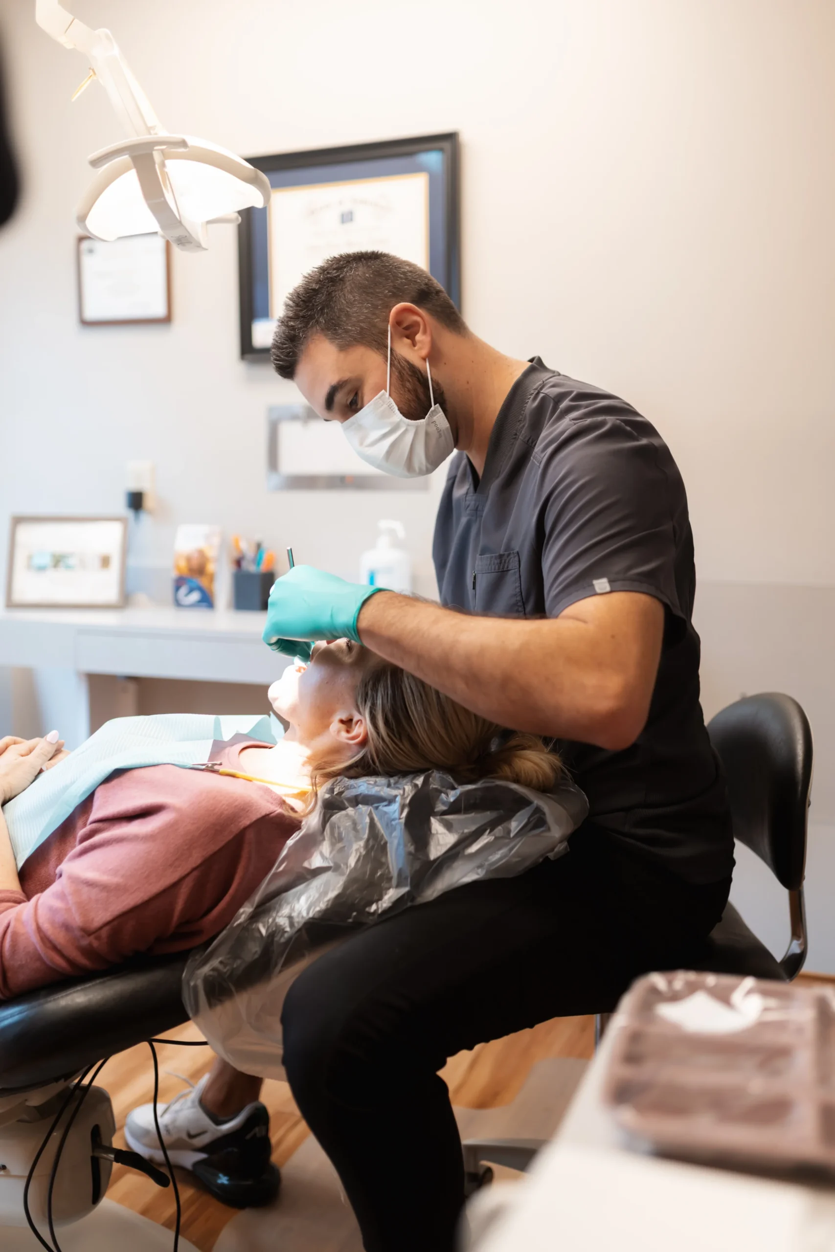 A dentist wearing a mask and gloves examines a patient reclining in a dental chair. The environment is clean, with certifications on the wall.