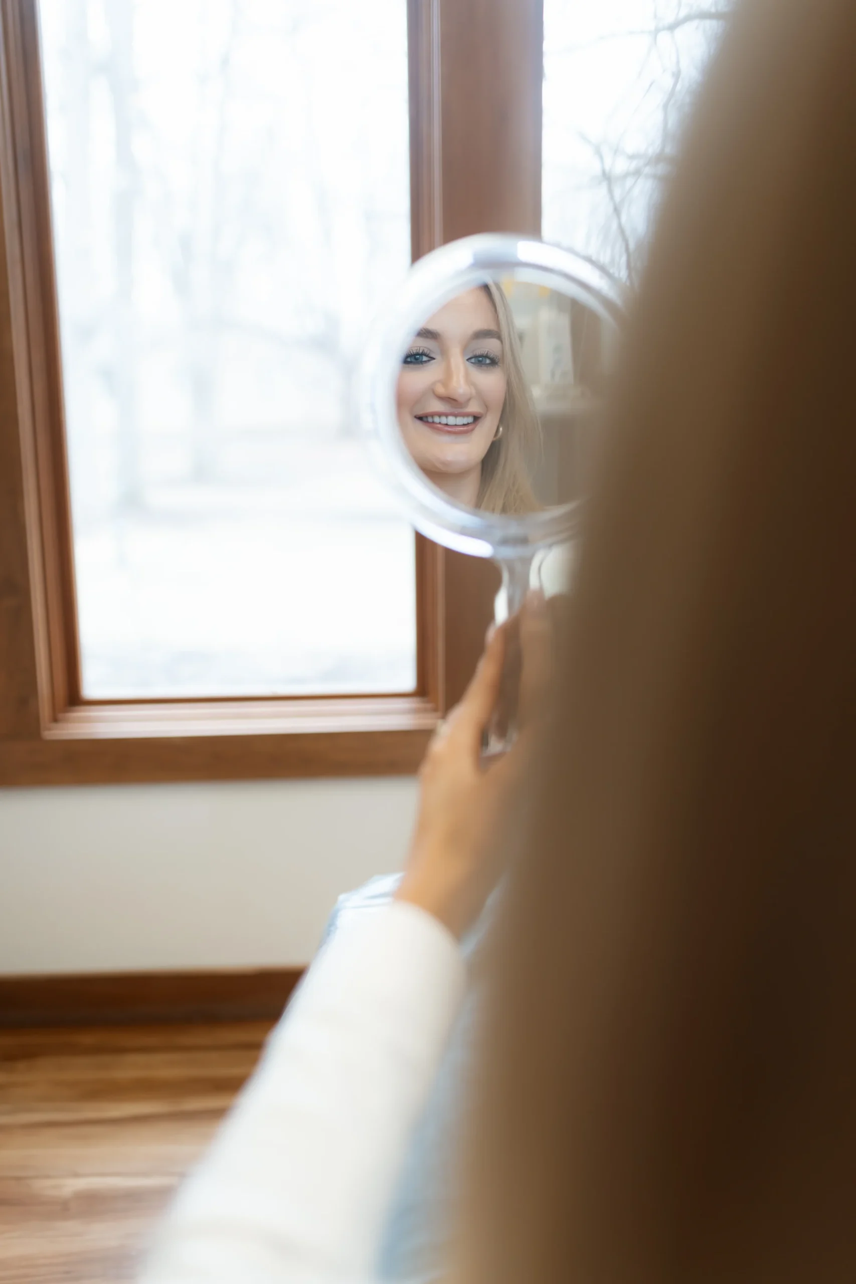 A woman smiles into a round mirror, her reflection showing bright eyes and long hair. The background is softly blurred, creating a joyful, serene atmosphere.